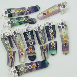 May include: A collection of 14 chakra pendants made from fluorite with silver accents. Each pendant features seven colored gemstones representing the seven chakras. The pendants are arranged in a random pattern on a white surface.