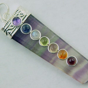 Puede incluir: Un colgante de plata con una piedra de fluorita púrpura, verde y blanca. El colgante tiene siete pequeñas piedras redondas de diferentes colores, que representan los siete chakras.