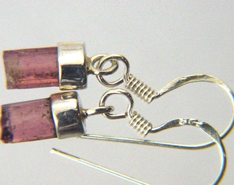 May include: A pair of silver earrings featuring pink tourmaline gemstones. The earrings have a simple design with a small, rectangular gemstone set in a silver setting. The earrings are attached to a silver hook.