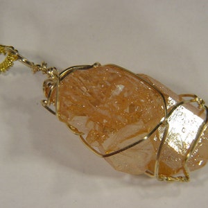 Può includere: Un ciondolo avvolto in filo d'oro con un grande cristallo grezzo color pesca.