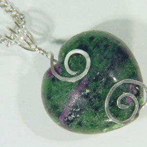 Op de afbeelding: Een groene hartvormige hanger met zilveren swirls, gemaakt van een groene en zwarte edelsteen. De hanger is bevestigd aan een zilveren ketting.