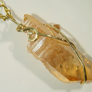 Op de afbeelding: Een grote, heldere, oranje kristallen hanger, omwikkeld met een gouden draad. De hanger is bevestigd aan een gouden ketting.