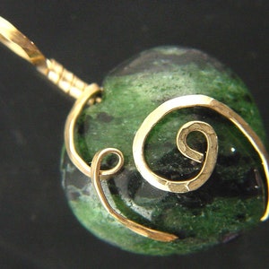 Op de afbeelding: Een groene en zwarte edelsteen hanger met een gouden draad eromheen gewikkeld in een swirl-ontwerp.