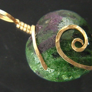 Op de afbeelding: Een groene en paarse edelsteen hanger met een gouden draadspiraal ontwerp. De hanger is bevestigd aan een gouden balein.