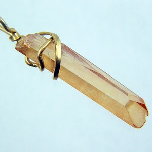 Op de afbeelding: Een lichtbruine kristallen hanger omwikkeld met gouden draad. De kristal is gefacetteerd en heeft een puntig uiteinde. De hanger is bevestigd aan een gouden balein.