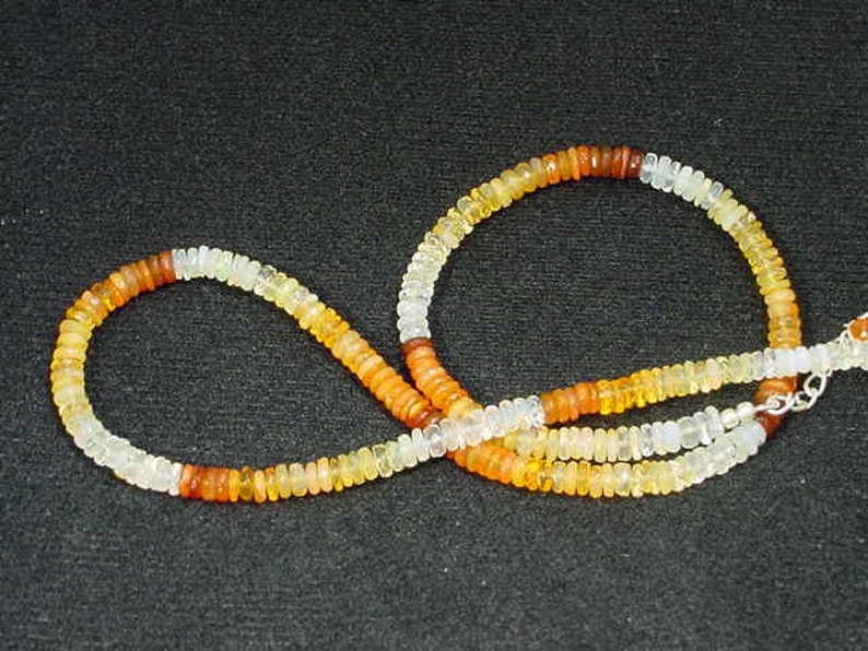 May include: A necklace made of small, round, multi-colored opal beads. The beads are arranged in a graduated pattern, with the darkest beads at the top and the lightest beads at the bottom. The necklace is strung on a silver chain.