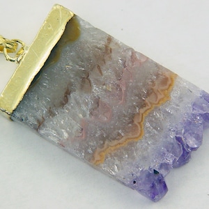 Può includere: Un ciondolo placcato oro con un cristallo di ametista grezzo con sfumature viola e bianche. Il cristallo ha una texture ruvida e naturale ed è incastonato in una cornice placcata oro.