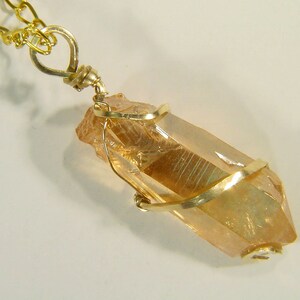 Op de afbeelding: Een gouden ketting met een hanger met een heldere, gefacetteerde, gouden kwartskristal gewikkeld in gouden draad.