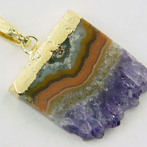 Può includere: Un ciondolo con bordo dorato che presenta un cristallo di ametista viola con una base di agata a strati. Il ciondolo ha un anello dorato per fissarlo a una collana.
