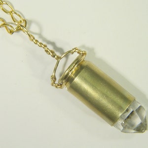 Può includere: Una collana con catena dorata con un ciondolo fatto di una punta di cristallo trasparente racchiusa in un bossolo di proiettile dorato.