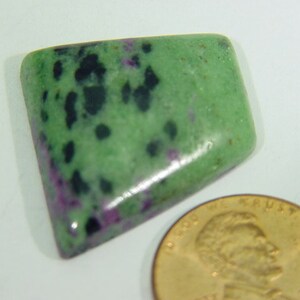 May include: A polished, rectangular gemstone with a green base color, speckled with black and flecks of purple. A gold-colored coin is next to the gemstone. The gemstone is likely a cabochon, used in jewelry making.