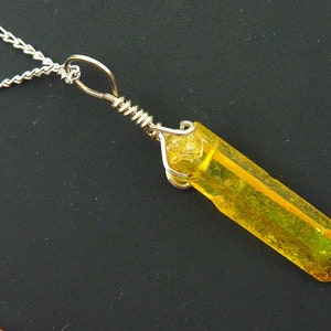 Op de afbeelding: Een zilveren ketting met een gele kristallen hanger. Het kristal heeft een lange, rechthoekige vorm met een puntig uiteinde en is omwikkeld met zilverdraad. De achtergrond is zwart.