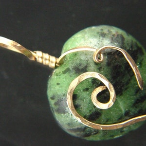 Op de afbeelding: Een groene en zwarte edelsteen hartvormige hanger met een gouden draad eromheen gewikkeld in een spiraalvorm.