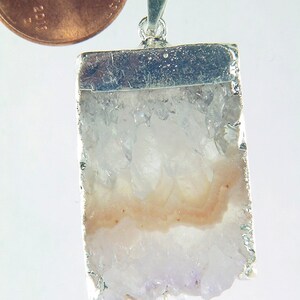Może przedstawiać: Srebrny wisiorek z przezroczystym kryształem kwarcu i różową wstążką. Wisiorek jest mniej więcej wielkości polskiego grosza.