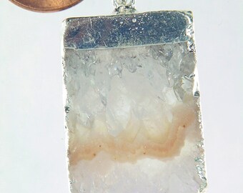Puede incluir: Un colgante de plata con un cristal de cuarzo transparente y una banda rosa. El colgante es aproximadamente del tamaño de una moneda de un céntimo de euro.
