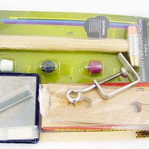 May include: A 5-in-1 interchangeable hammer set with a wooden handle, a metal hammer head, and a variety of interchangeable tips. The set also includes a clamp, a wooden block with holes, and a small metal tool. The packaging is green and yellow with the text "5-IN-1 INTERCHANGEABLE HAMMER 8355HH 9" printed on it.