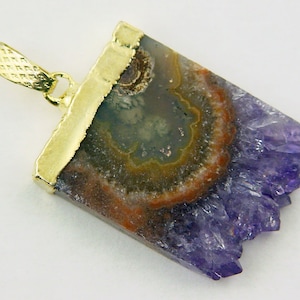 Può includere: Un ciondolo placcato oro con un cristallo di ametista viola e un centro di agata marrone e arancio. Il cristallo ha una texture ruvida e naturale ed è a forma di rettangolo con un bordo appuntito.