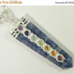 Puede incluir: Un collar de cadena de plata con un colgante de piedra azul. El colgante tiene siete pequeñas piedras redondas de diferentes colores engastadas en plata a lo largo del lado. Las piedras representan los siete chakras.