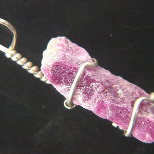 Könnte beinhalten: Ein Anhänger aus rohem rosa Rubin-Edelstein, der in Silberdraht gewickelt ist. Der Anhänger ist an einer silbernen Kette mit einem Perlendetail befestigt.