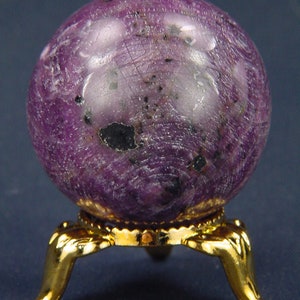 May include: A purple gemstone sphere on a gold metal stand. The sphere has a rough, textured surface with black flecks throughout.