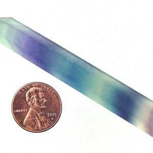 Puede incluir: Una varita de cristal de fluorita larga y puntiaguda, de color verde, azul y morado. El cristal mide aproximadamente 7,5 cm de largo y está al lado de un centavo estadounidense para la escala.