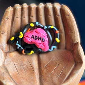 May include: A handcrafted sculpture featuring a pink brain with the letters "ADHD" and a black, swirled design with colorful accents. The sculpture rests in a carved wooden hand, showcasing a unique artistic piece.
