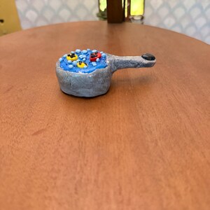 May include: A small, hand-sculpted gray pan with a textured finish, filled with a bright blue substance resembling liquid. The surface features small, raised yellow and red circular shapes, along with tiny white dots scattered throughout.
