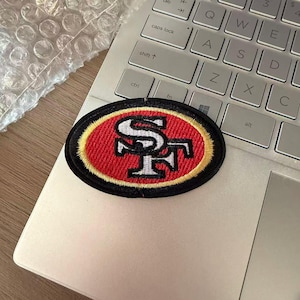 Puede incluir: Un parche bordado de forma ovalada con el logotipo de los San Francisco 49ers. El diseño incluye un fondo rojo con un borde dorado y las letras SF en blanco con contornos negros, todo ello encerrado en un grueso borde negro cosido.