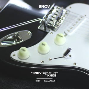May include: An electric guitar body featuring a black finish, a white pickguard, and three off-white control knobs labeled VOLUME and TONE. The text 8NOV is visible at the top, with 8NOV signature KNOB and 8nov_official printed at the bottom.