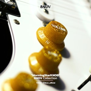 Puede incluir: Tres perillas de volumen de guitarra con purpurina dorada y marcas numéricas del 1 al 10, montadas en un cuerpo de guitarra eléctrica blanco. La perilla superior está enfocada con la palabra VOLUME impresa. Texto: 8NOV, MorningStarKNOB, 8NOV Collection.