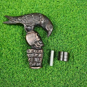 May include: A bronze-toned metal cane handle featuring a sculpted eagle perched above a human skull, which rests on a rectangular base with a grid pattern. The set includes a threaded metal bolt and a cylindrical metal collar attachment.