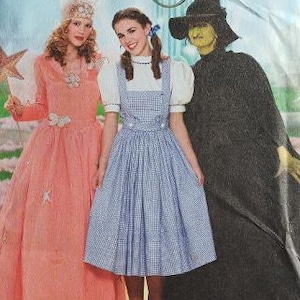 May include: A Simplicity sewing pattern envelope for The Wizard of Oz costumes. The front shows three outfits: a long peach gown with star accents, a blue and white gingham pinafore dress with a white blouse, and a long black hooded cloak.