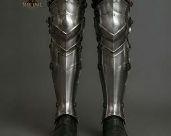 Könnte beinhalten: Ein Paar Beinschienen aus poliertem Stahl, die Knie und Schienbeine bedecken, mit beweglichen Metallplatten, Nieten und Lederriemen zur sicheren Befestigung. Die Rüstung wird über einer dunklen Hose und schwarzen Lederschuhen getragen.