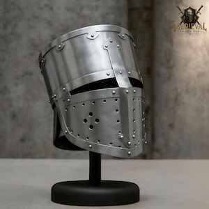 Puede incluir: Un casco medieval plateado con un diseño remachado, con una visera con múltiples agujeros pequeños. El casco se exhibe sobre un soporte negro, mostrando su artesanía y diseño históricos. El casco es de metal.