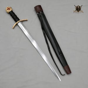 May include: A steel sword with a double-edged blade, a black leather-wrapped grip, and a gold-toned crossguard. The pommel features a gold-toned cross emblem. Beside the sword lies a black leather scabbard with a brown leather tip and straps.