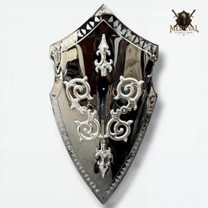 May include: A polished silver shield with ornate, scrolling silver embellishments. The shield has a pointed base and a decorative border. The shield is reflective, with the Medieval Armor Hub logo visible in the upper right corner.