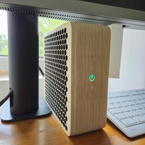 Puede incluir: Una pequeña torre de ordenador de color claro con un panel de malla de panal y un panel lateral de vetas de madera verticales. La torre tiene un indicador de encendido verde y está sobre un escritorio de madera cerca de un teclado y un monitor.