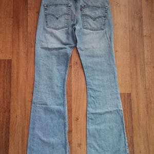 May include: Light blue denim jeans with a flared leg design. The jeans feature classic five-pocket styling, a button and zipper closure, and a leather patch on the back. The back pockets have a signature curved stitch.
