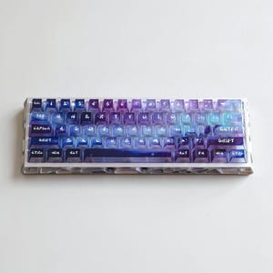 May include: A mechanical keyboard featuring a gradient colour scheme transitioning from deep purple to light blue. The keys are translucent with white lettering in a handwritten font style. The keyboard frame has a metallic, geometric, open-work design.
