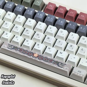 Könnte beinhalten: Eine mechanische Tastatur mit einem mehrfarbigen Tastenkappenset in Weiß, Dunkelgrau und Kastanienbraun. Die weißen Tasten zeigen schwarze Buchstaben und florale Symbole. Die Leertaste ist mit einem komplexen, dekorativen Rankenmuster verziert.