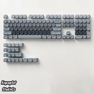 Peut inclure: Un ensemble complet de touches de clavier mécanique arrondies dans des nuances de gris variées. La disposition comprend un agencement QWERTY standard, un pavé numérique, des touches de fonction et des touches modificatrices supplémentaires.