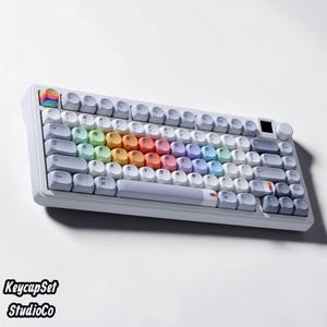 May include: A white mechanical keyboard featuring rounded keycaps. The central keys display a gradient of pastel colors including green, yellow, orange, pink, and blue. The escape key and spacebar feature small rainbow accents. A volume knob is present.
