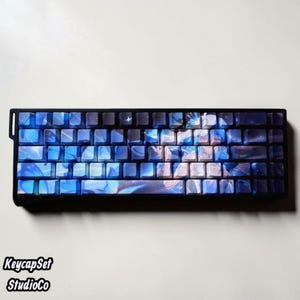 Könnte beinhalten: Eine mechanische Tastatur mit einem individuellen Tastenkappen-Set in Blau und Schwarz. Die Tasten zeigen eine aufgedruckte Grafik einer Anime-Figur, umgeben von blauen Schmetterlingen und funkelnden Lichteffekten auf der gesamten Oberfläche.