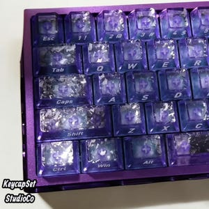 Puede incluir: Primer plano de un teclado mecánico morado con teclas transparentes. Las teclas tienen una apariencia cristalina y están dispuestas en una distribución QWERTY estándar. El marco del teclado es de color morado liso. El texto "KeycapSet StudioCo" es visible.