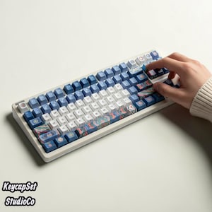 May include: A mechanical keyboard featuring a white frame and a custom keycap set with a mix of solid blue, white, and patterned keys. The patterned keys display intricate, colourful wave-like designs in shades of blue, orange, and teal.