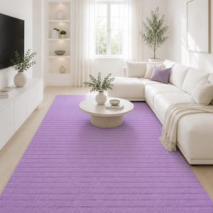 May include: A large, rectangular, lilac rug with a ribbed texture in a living room setting. The rug is placed in front of a white sectional sofa and a round coffee table. The room has white walls and a large window.