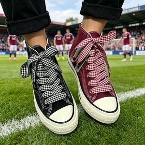 May include: A pair of high-top canvas sneakers, one black and one burgundy, with white rubber soles and toe caps. Each shoe features gingham patterned laces tied in a bow. The shoes are on a green field.