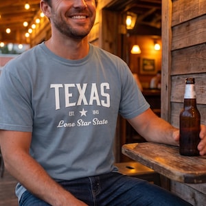 Puede incluir: Camiseta azul claro con la palabra "TEXAS" en blanco, una estrella y el texto "Lone Star State". Una botella de cerveza marrón con una etiqueta plateada se sostiene cerca de una mesa de madera.