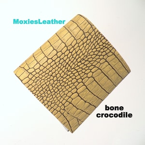 May include: A close-up of a piece of bone-colored crocodile embossed leather. The leather has a textured surface with a pattern of scales. The text "MoxiesLeather" is visible in the top left corner of the image. The text "bone crocodile" is visible in the bottom right corner of the image.