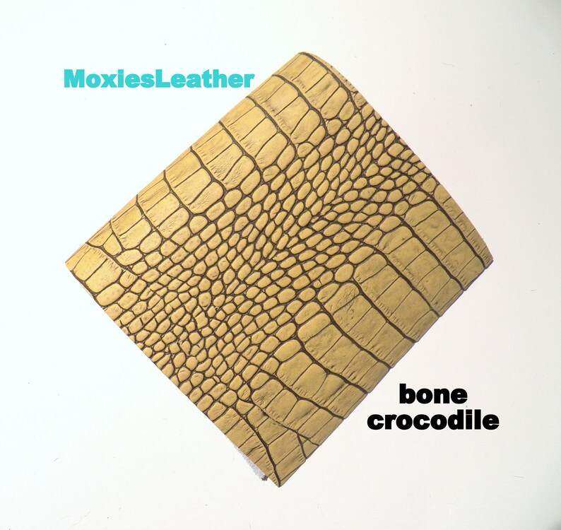 May include: A close-up of a piece of bone-colored crocodile embossed leather. The leather has a textured surface with a pattern of scales. The text "MoxiesLeather" is visible in the top left corner of the image. The text "bone crocodile" is visible in the bottom right corner of the image.